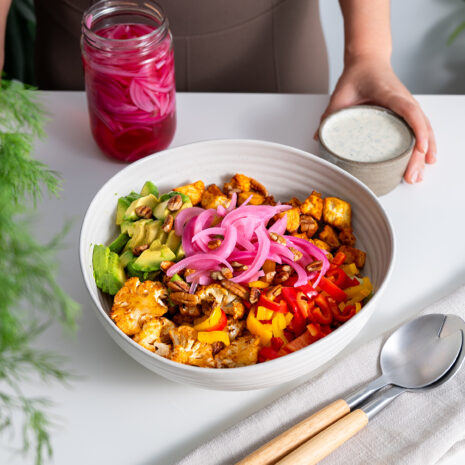 Full view of buffalo cauliflower salad with avocado and a tangy, herb-infused ranch dressing with Sarah in the background holding the dressing.