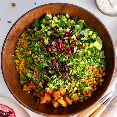 Overhead shot of a wooden bowl filled with a colorful couscous pomegranate salad on a white table.