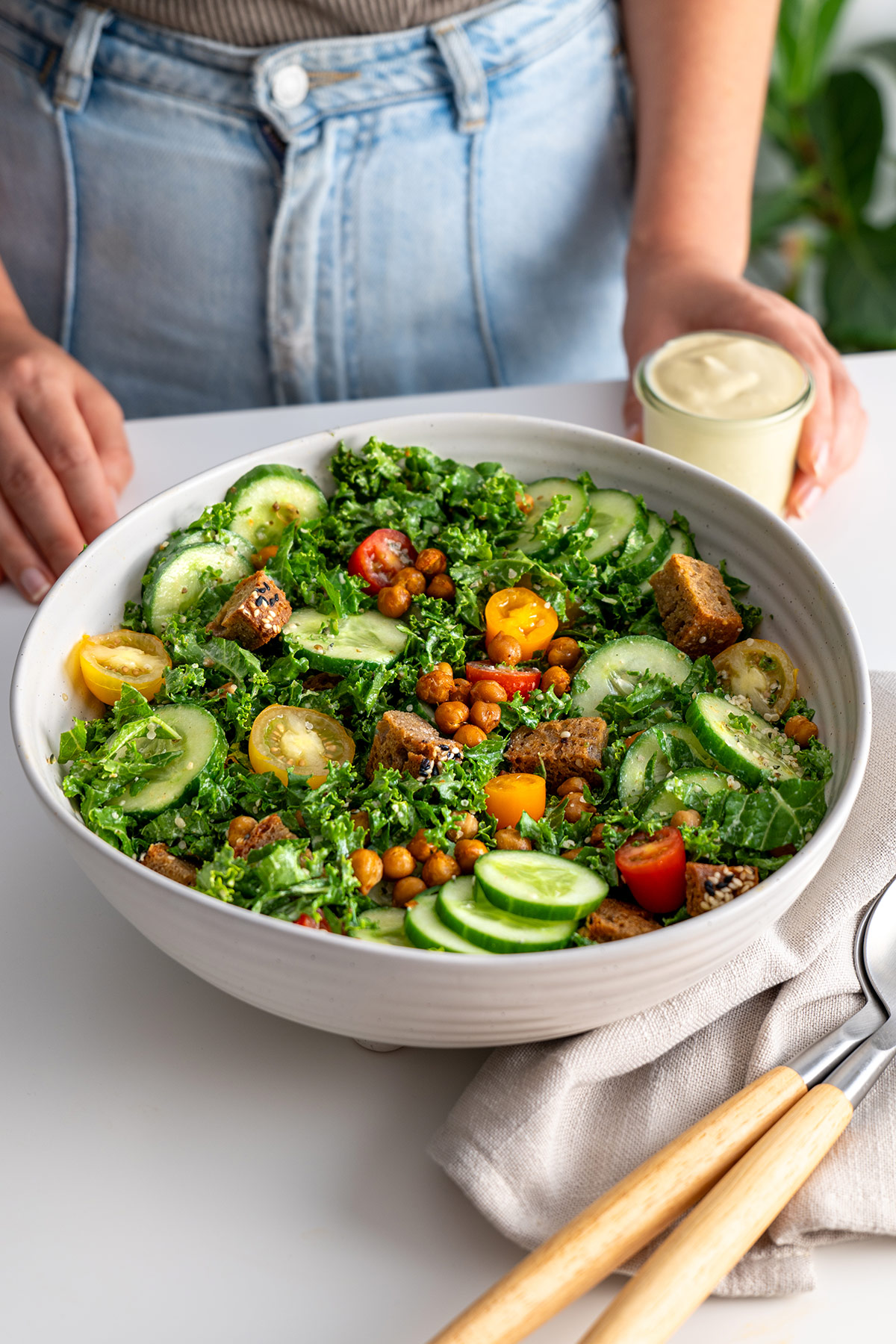 Hands holding a bowl of High-Protein Kale Caesar Salad with a creamy dressing on the side.