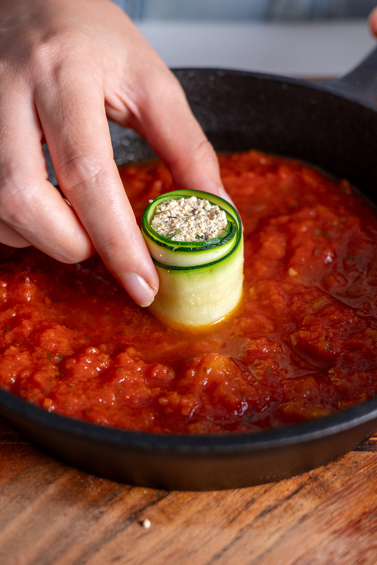 Close-up of a hand placing a Mediterranean zucchini roll-up filled with tofu mixture into a skillet with tomato sauce.