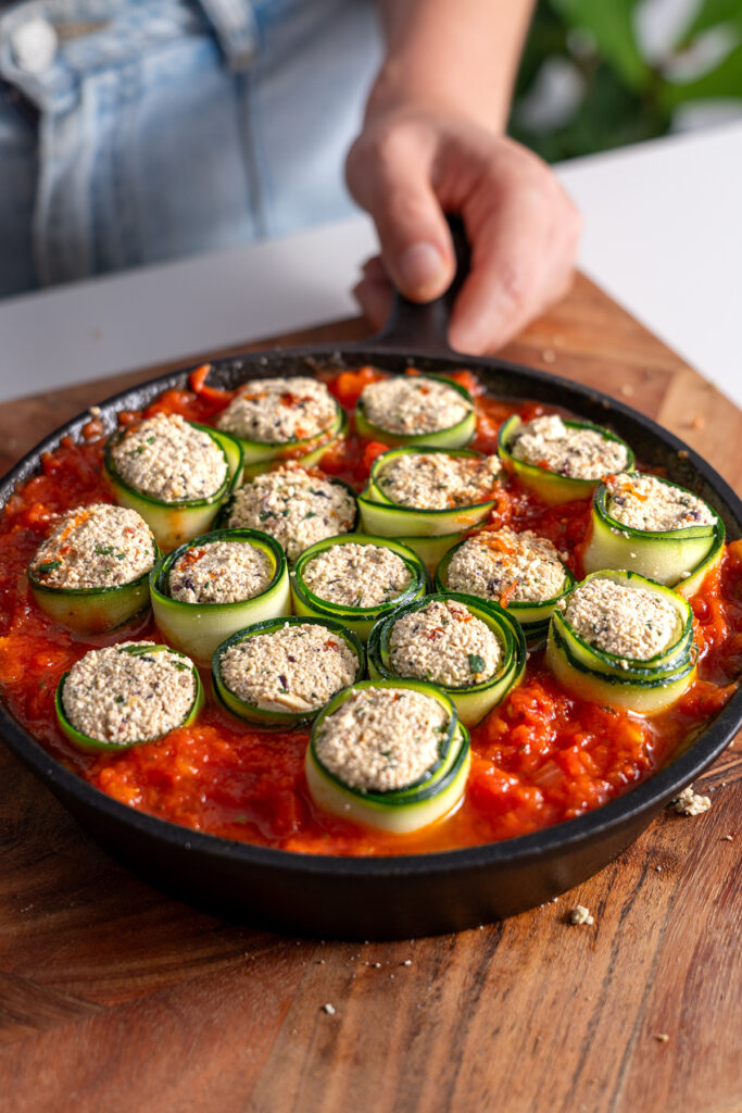 Hand placing a cast iron skillet of Mediterranean zucchini roll-ups ready to be baked on a wooden surface.
