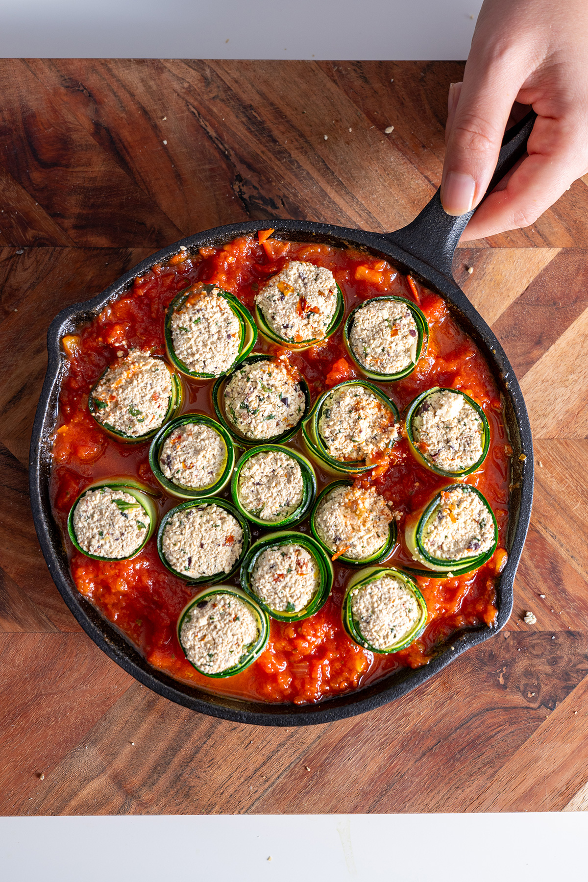 Hand placing a cast iron skillet of Mediterranean zucchini roll-ups ready to be baked on a wooden surface.