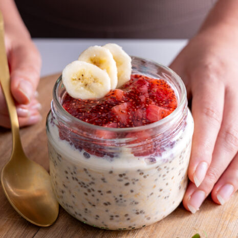 A hand holding a glass jar of overnight oats topped with strawberry jam and banana slices.