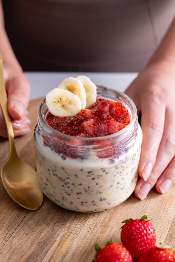 A hand holding a glass jar of overnight oats topped with strawberry jam and banana slices.