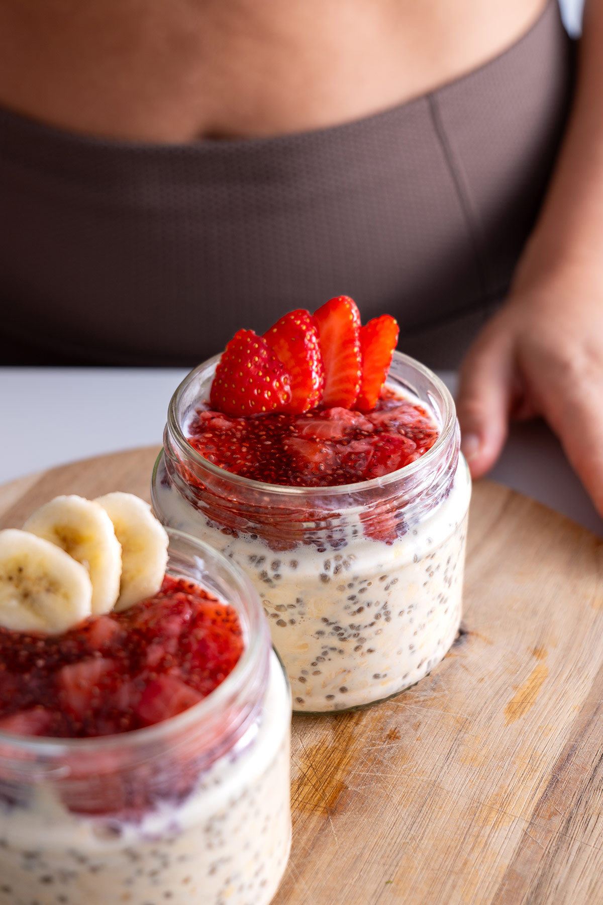 Glass jars of overnight oats layered with strawberry jam, garnished with banana slices and a strawberry fan.