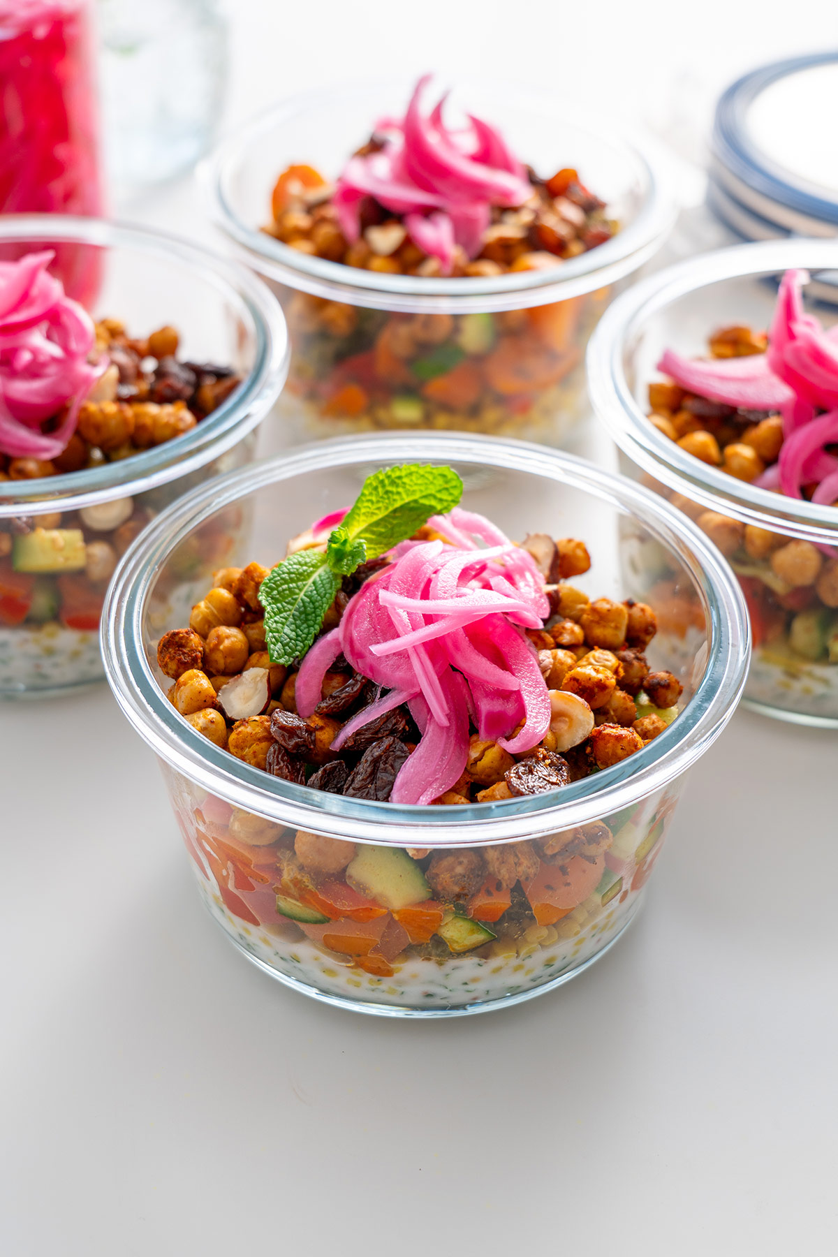 Glass meal prep containers filled with couscous salad, roasted chickpeas, and garnished with mint leaves and pickled onions.