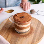 Tiramisu overnight oats in a clear glass on a wooden serving board with a spoon on the side.