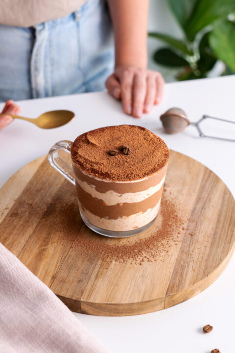 Tiramisu overnight oats in a clear glass on a wooden serving board with a spoon on the side.