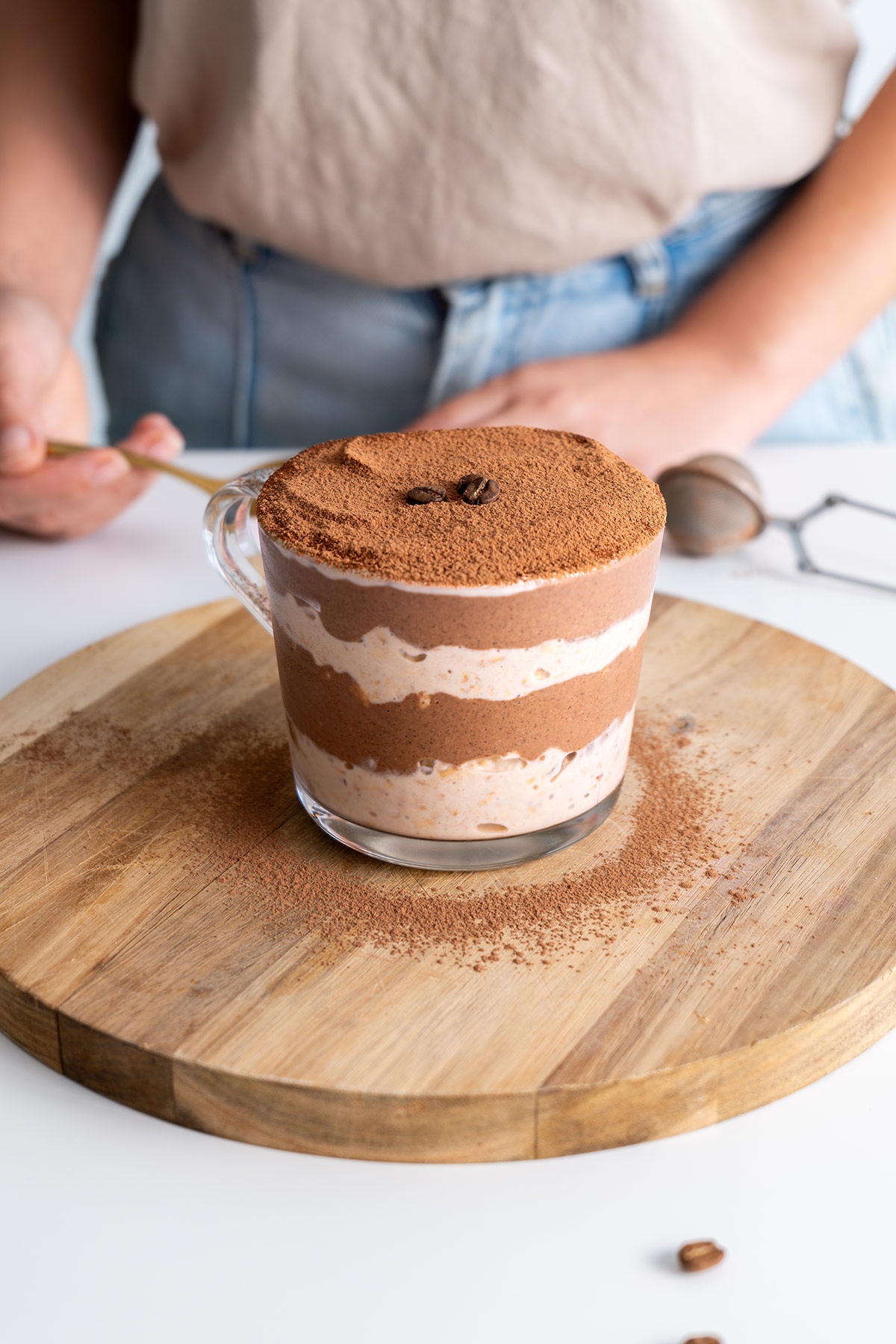 Close-up of tiramisu overnight oats in a glass jar Sarah Cobacho in the background holding a gold spoon.