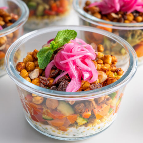 Close-up of a glass meal prep container with layers of dressing, roasted veggies and chickpeas with pickled onions on top.