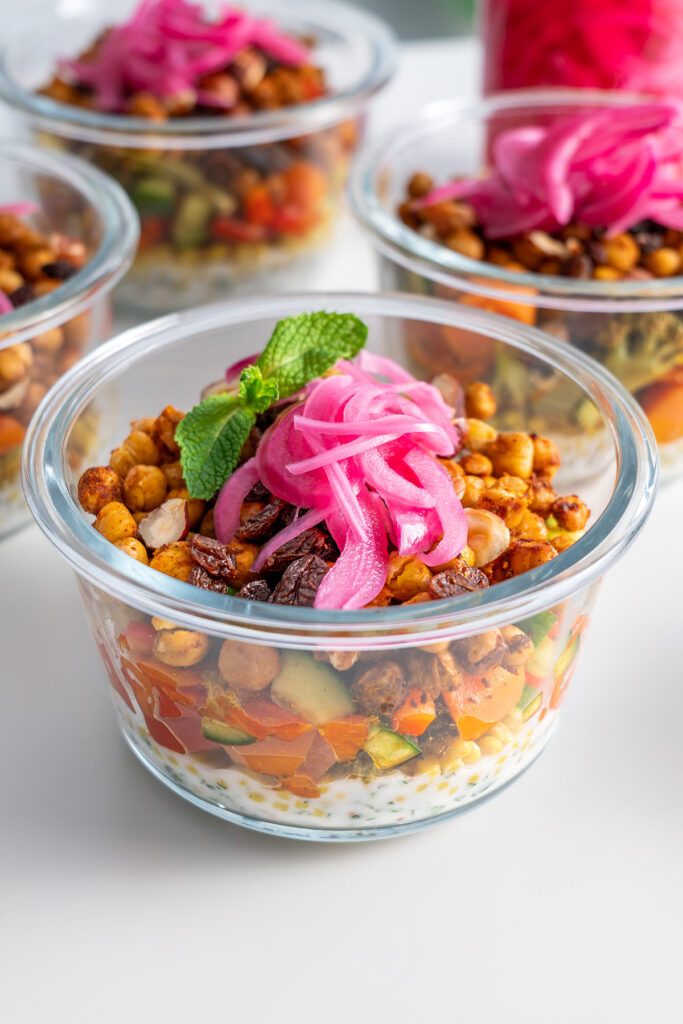 Close-up of a glass meal prep container with layers of dressing, roasted veggies and chickpeas with pickled onions on top.