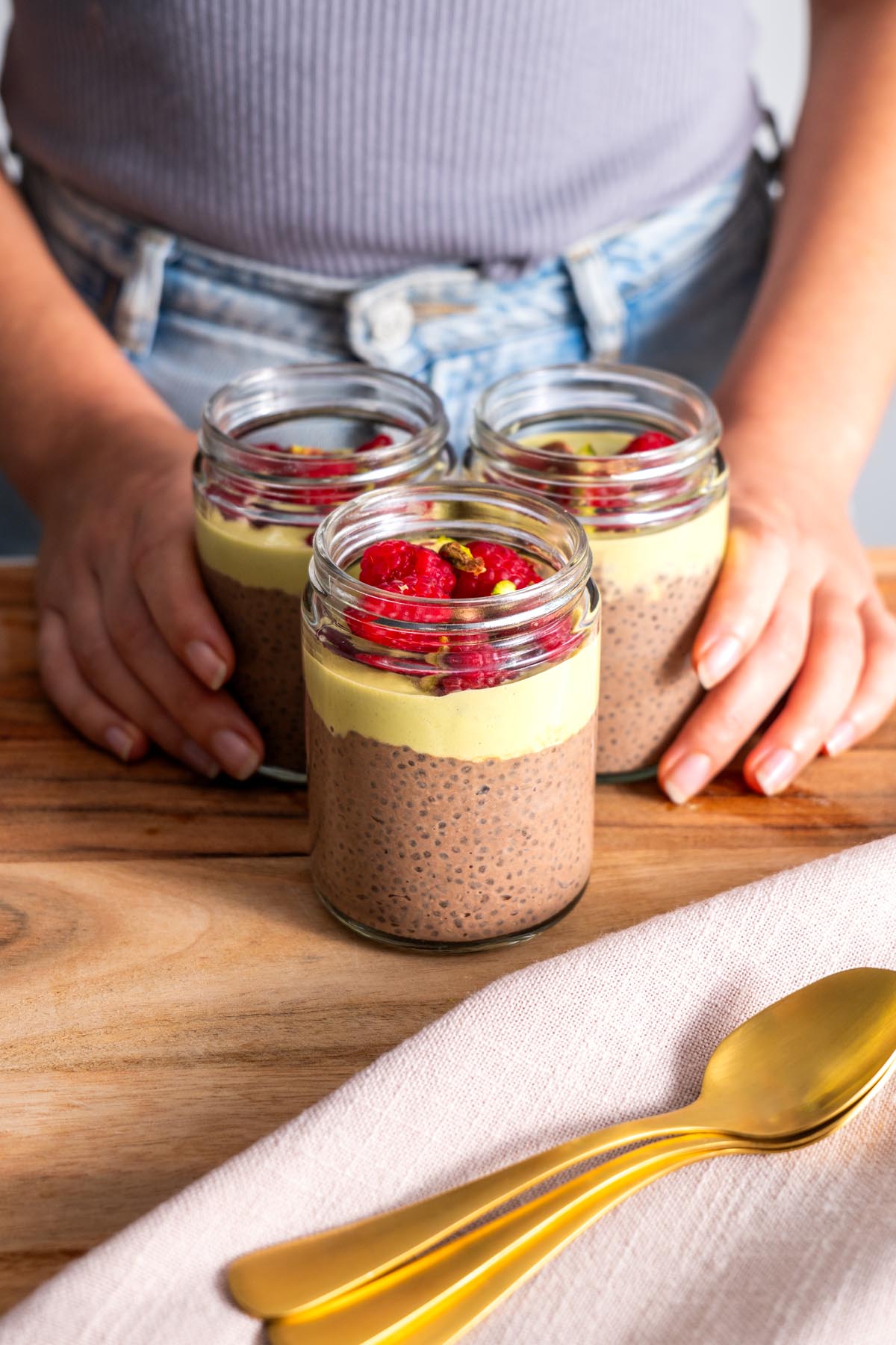 Three jars of Chocolate Pistachio Chia Pudding with raspberry and pistachio toppings on a wooden board.