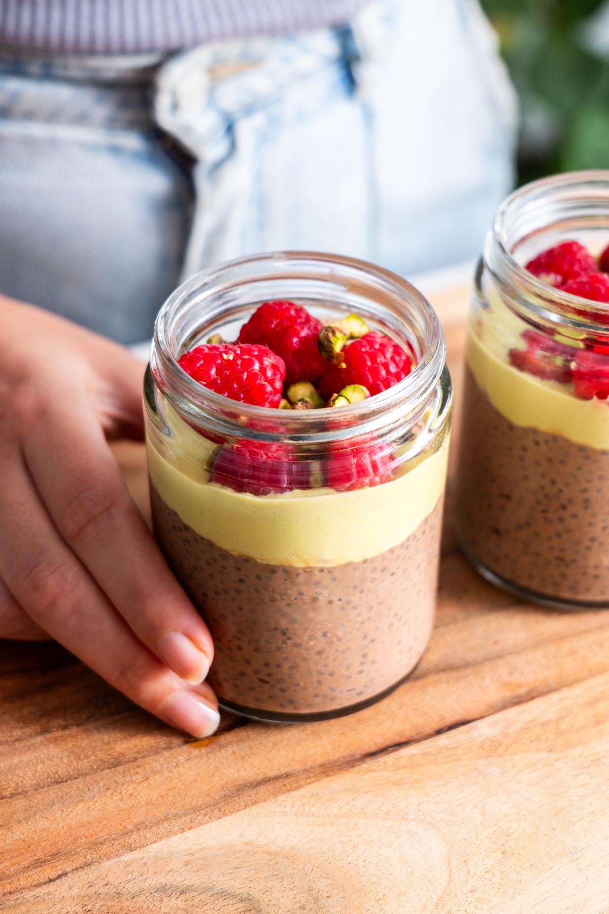 Sarah Cobacho preparing Chocolate Pistachio Chia Pudding in glass jars on a wooden surface.