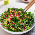 A large bowl of grilled broccolini and pomegranate salad with rocket and couscous on a white table, complete with serving utensils and fresh ingredients in the background.