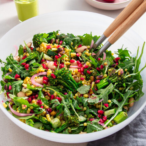 A large bowl of grilled broccolini and pomegranate salad with rocket and couscous on a white table, complete with serving utensils and fresh ingredients in the background.