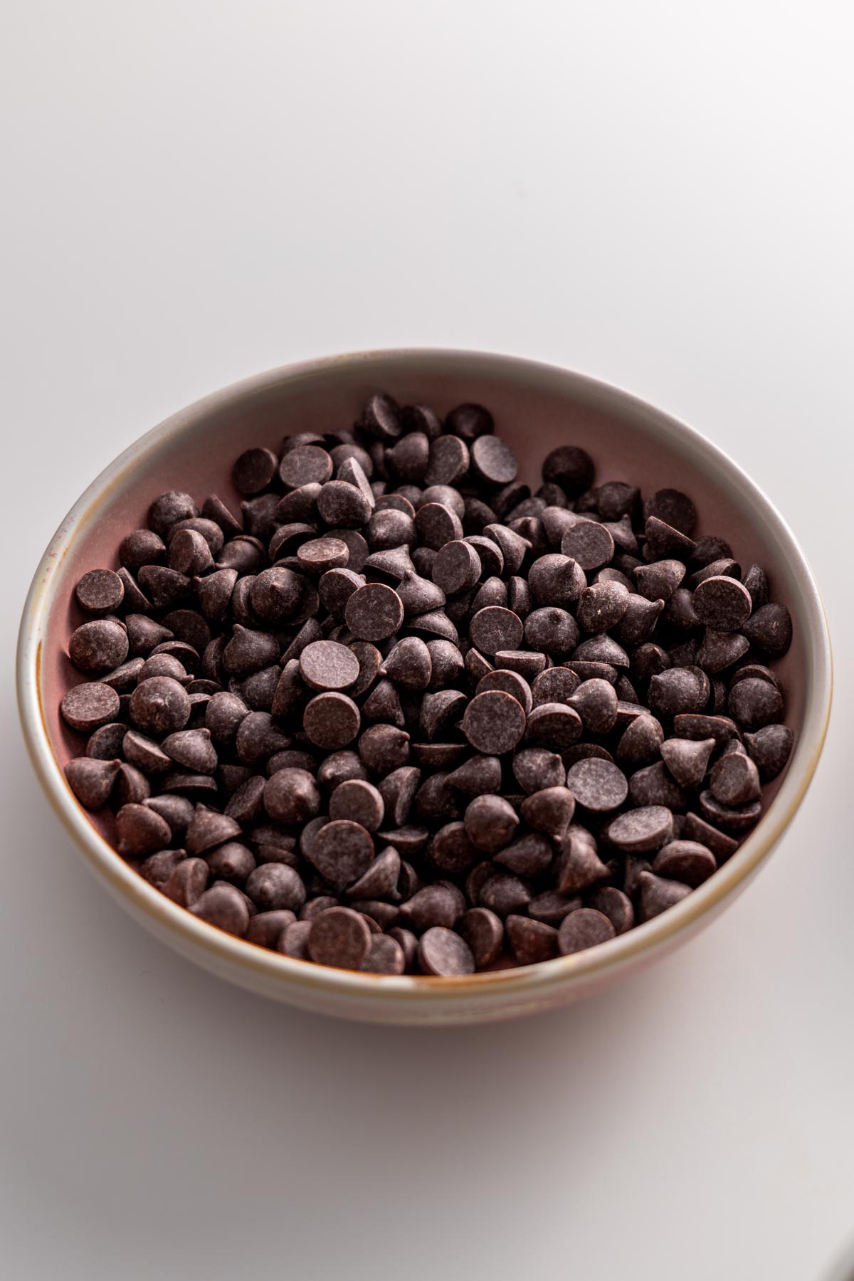 Dark chocolate chips in a light pink ceramic bowl on a white surface.