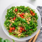 Top view of freshly prepared Pesto Orzo Salad in a white bowl on a kitchen counter, featuring arugula, broccolini, and cherry tomatoes.