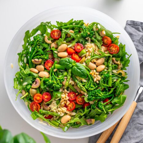 Top view of freshly prepared Pesto Orzo Salad in a white bowl on a kitchen counter, featuring arugula, broccolini, and cherry tomatoes.