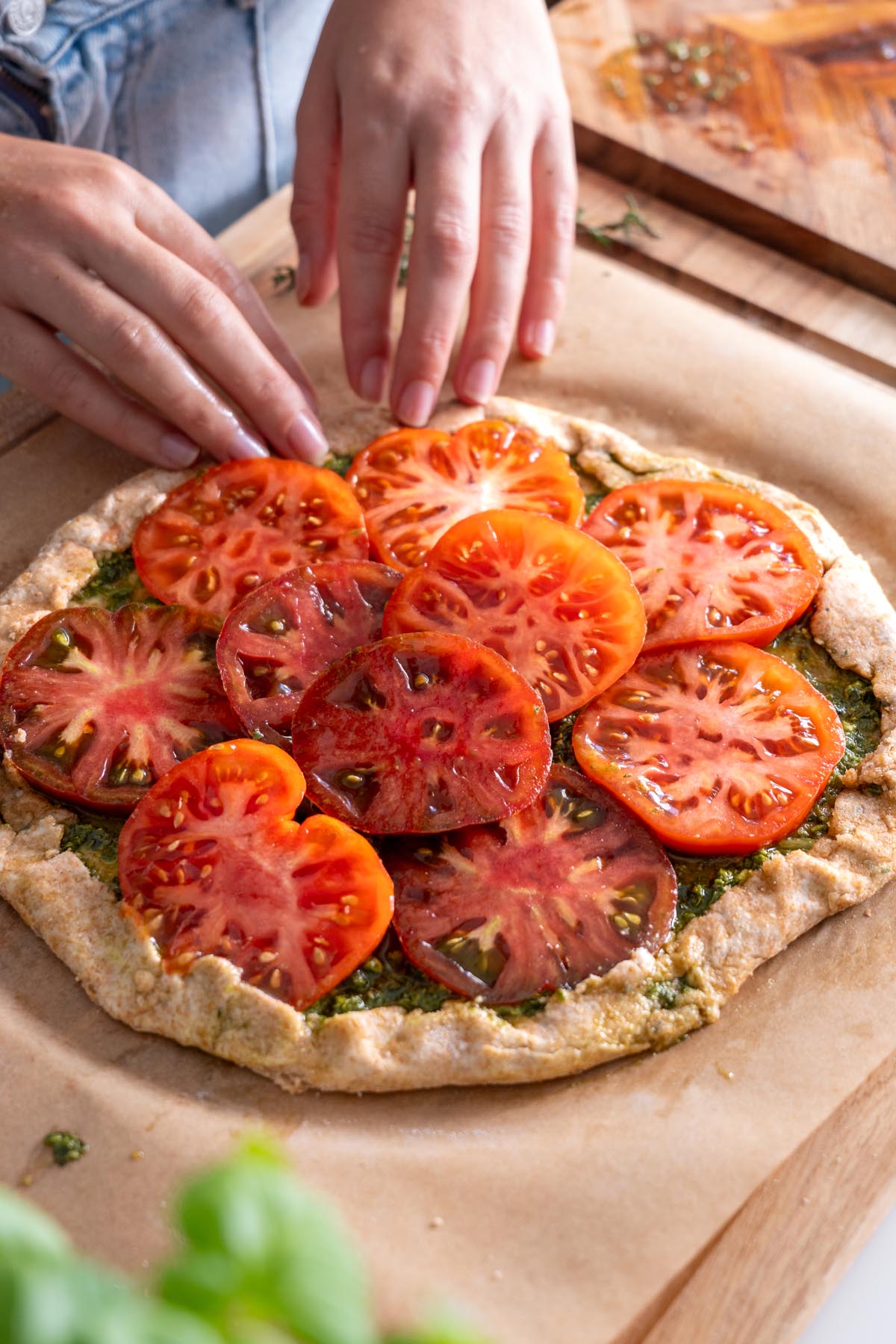 Hands arranging heirloom tomato slices on a galette dough spread with pesto.