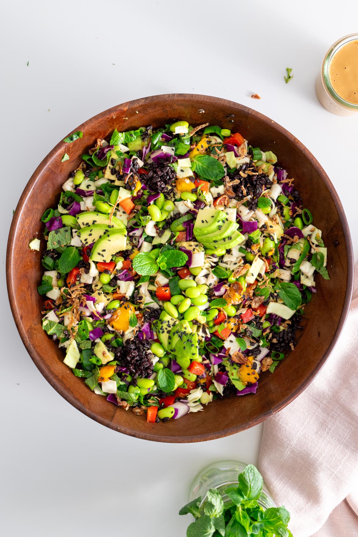 Top down view of rainbow salad with avocado, edamame, mango, and various fresh vegetables in a wooden bowl on a white surface.