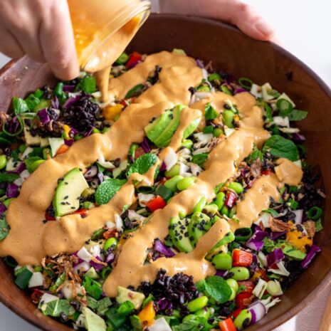 Pouring peanut dressing over a vibrant salad with a mix of fresh vegetables in a wooden bowl.