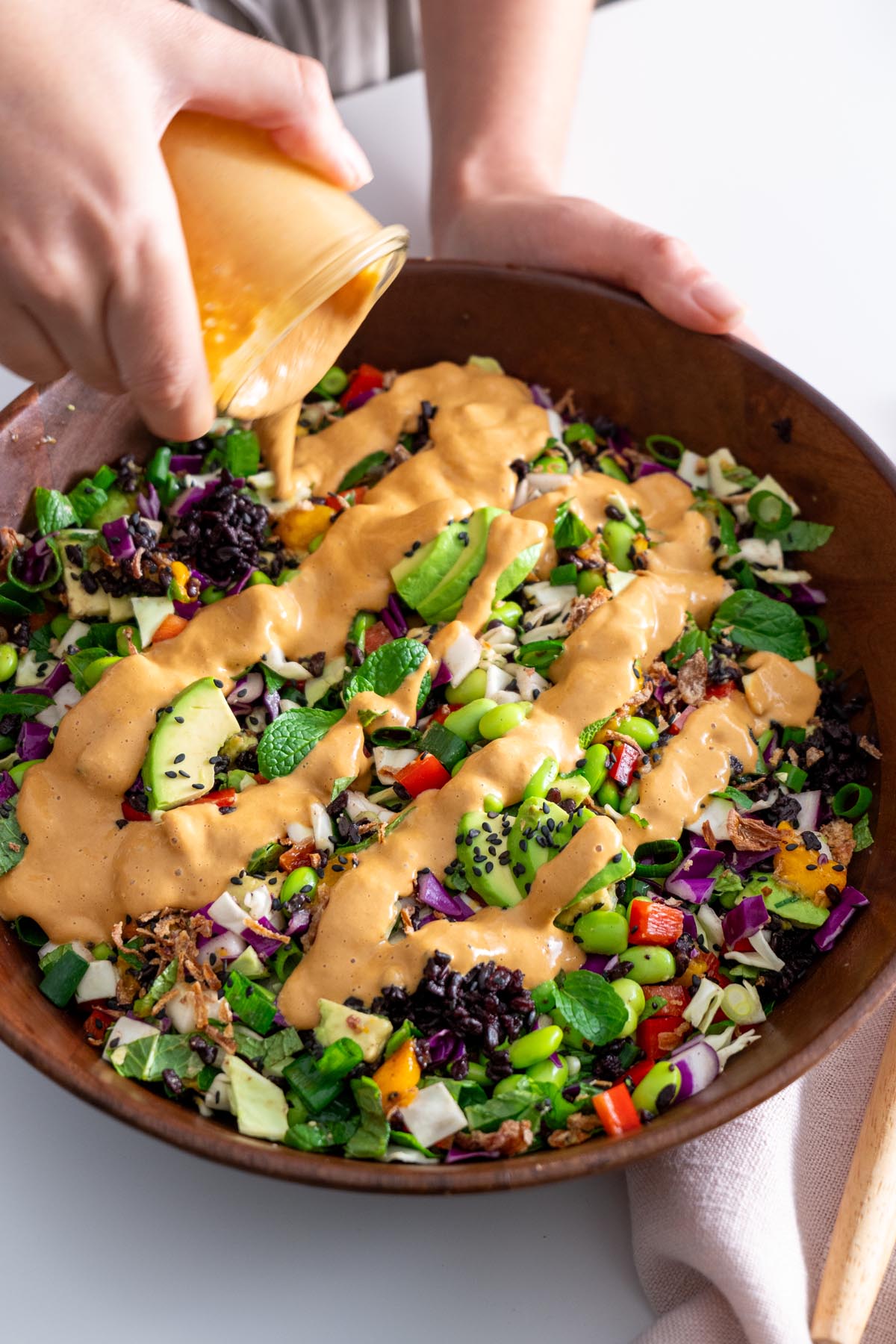 Pouring peanut dressing over a vibrant salad with a mix of fresh vegetables in a wooden bowl.