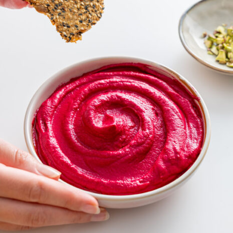 Smooth beetroot hummus in a white bowl, being served with a seed cracker.