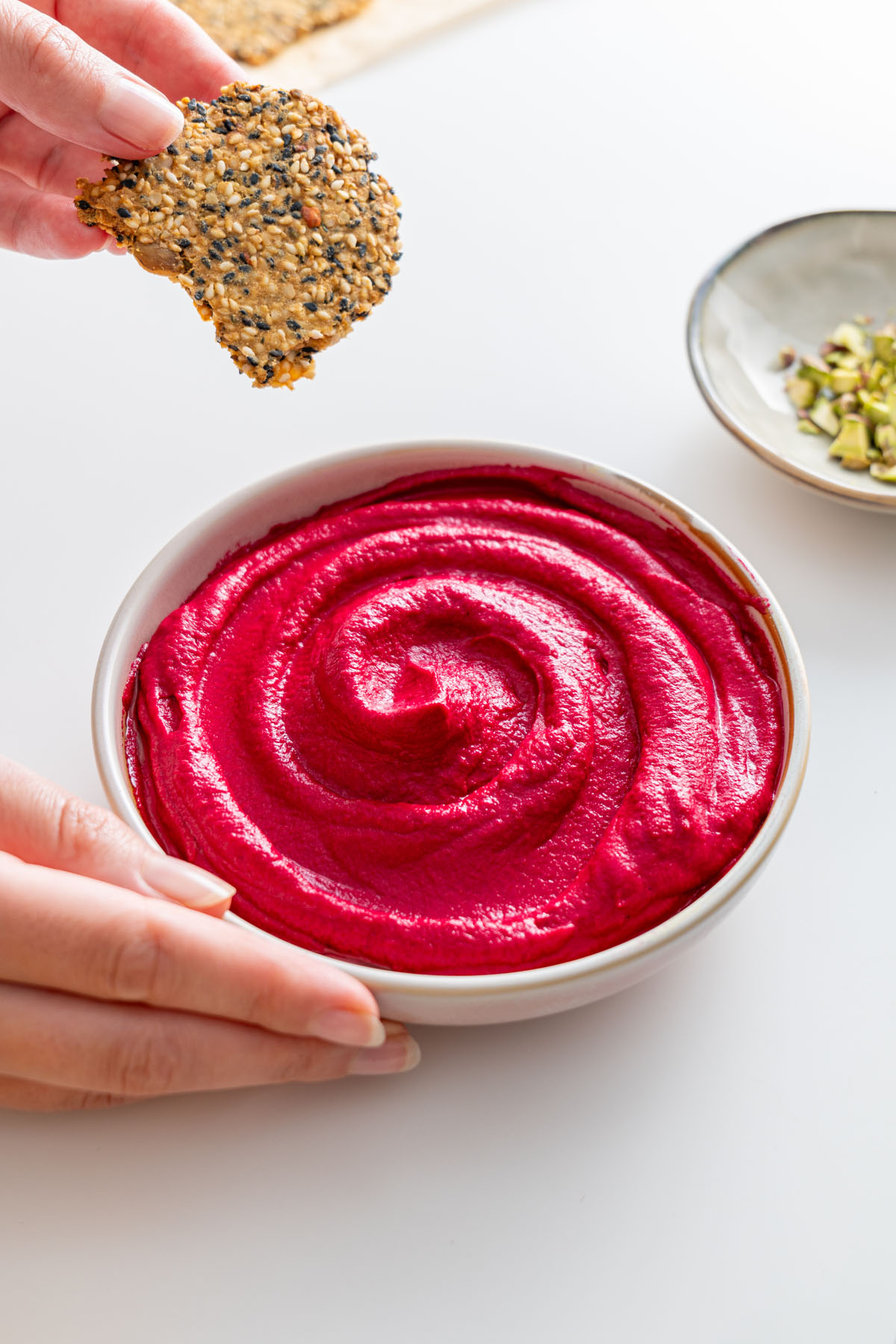 Smooth beetroot hummus in a white bowl, being served with a seed cracker.