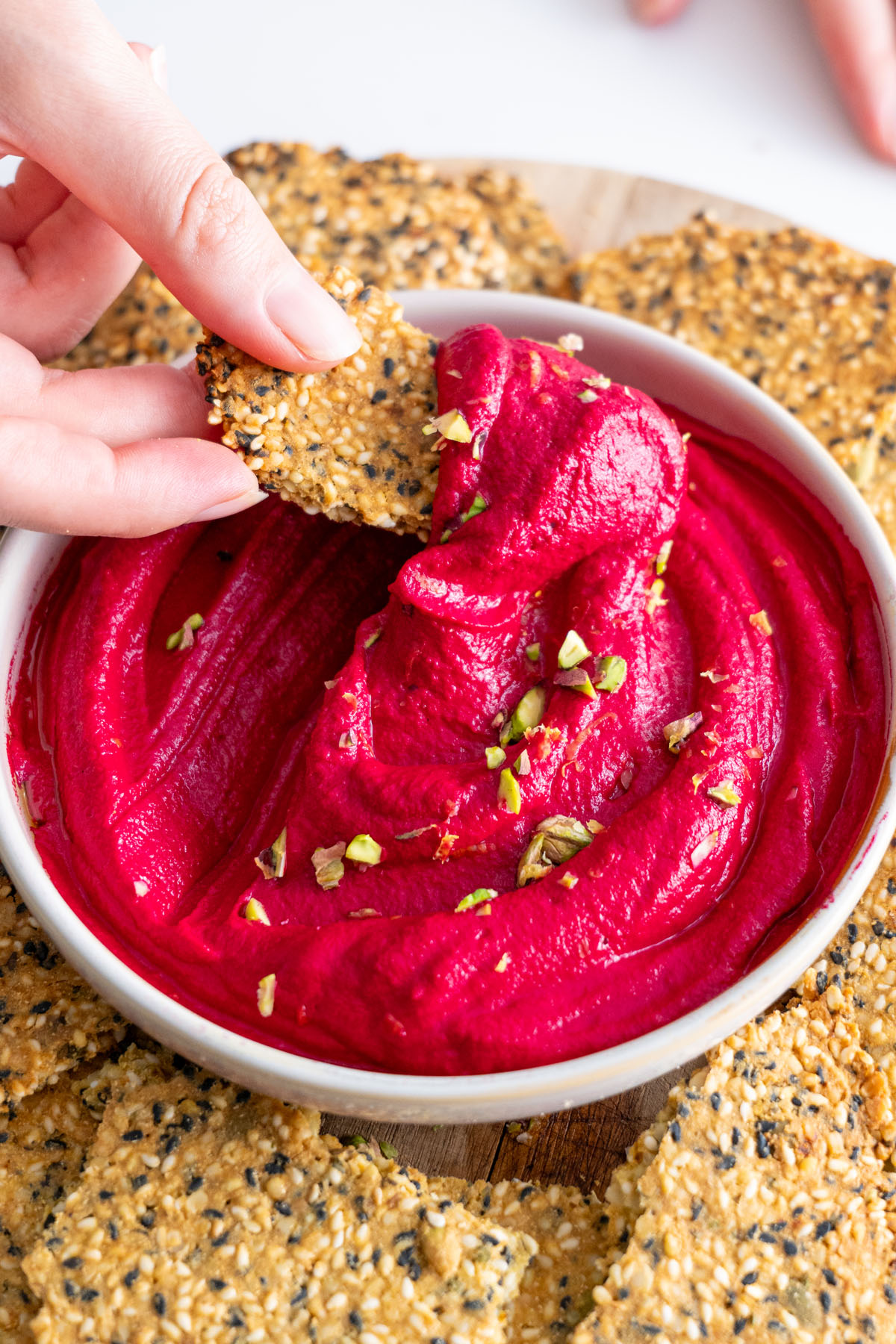Sarah's hand dipping a seed cracker into beetroot hummus topped with pistachios.