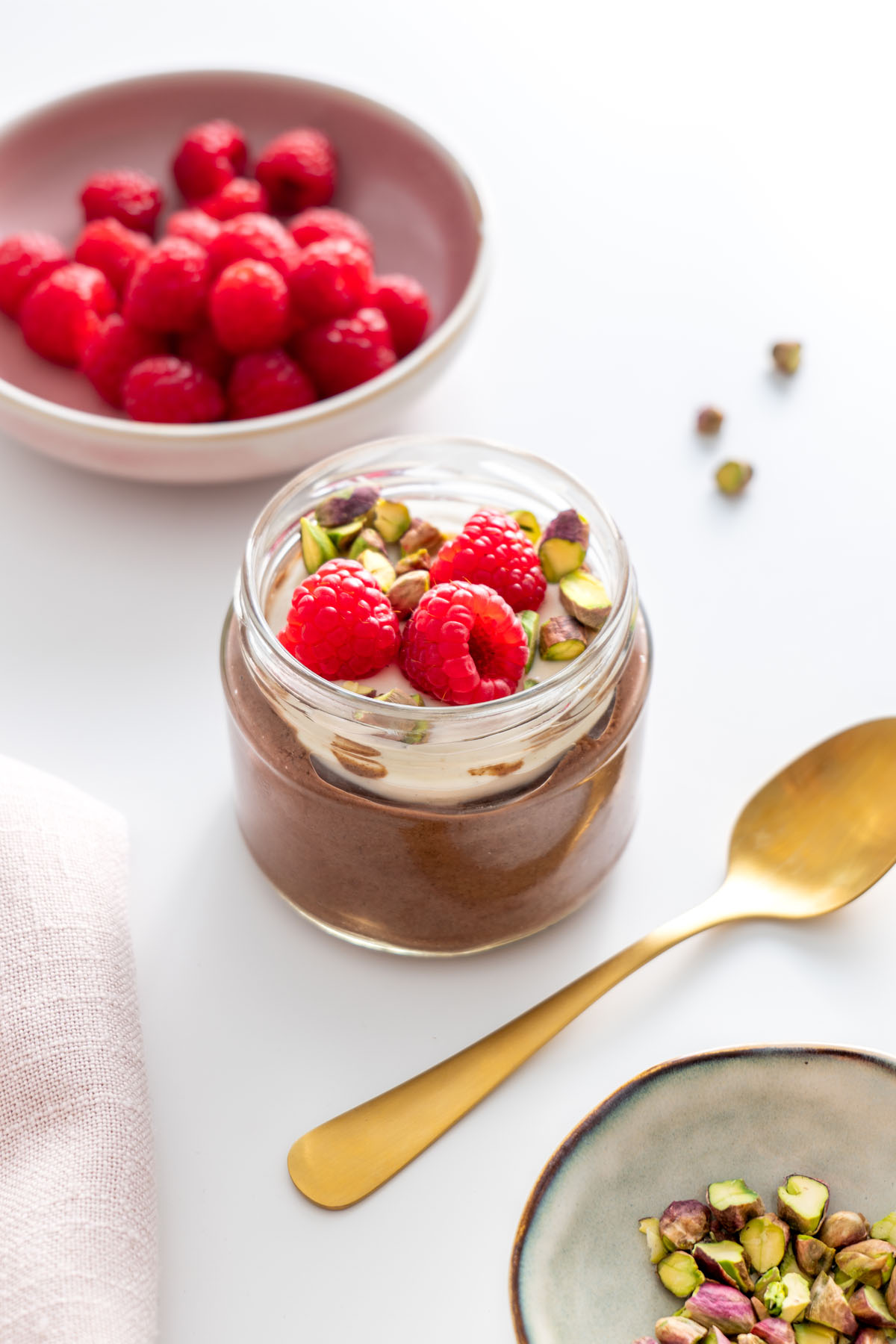 Cacao chia pudding jar topped with fresh raspberries and pistachios, with a spoon nearby.