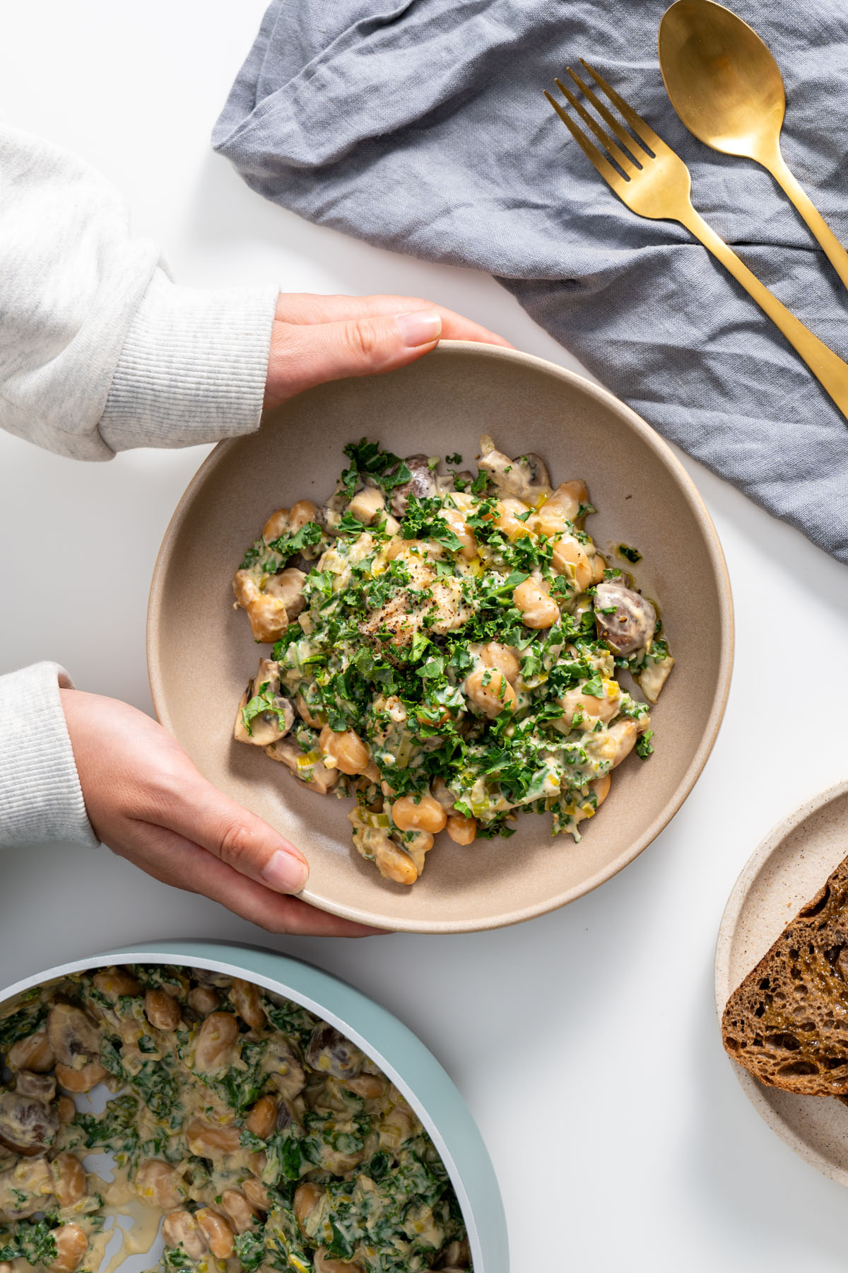 Sarah Cobacho’s hands holding creamy miso butter beans with kale next to toasted bread on a table.