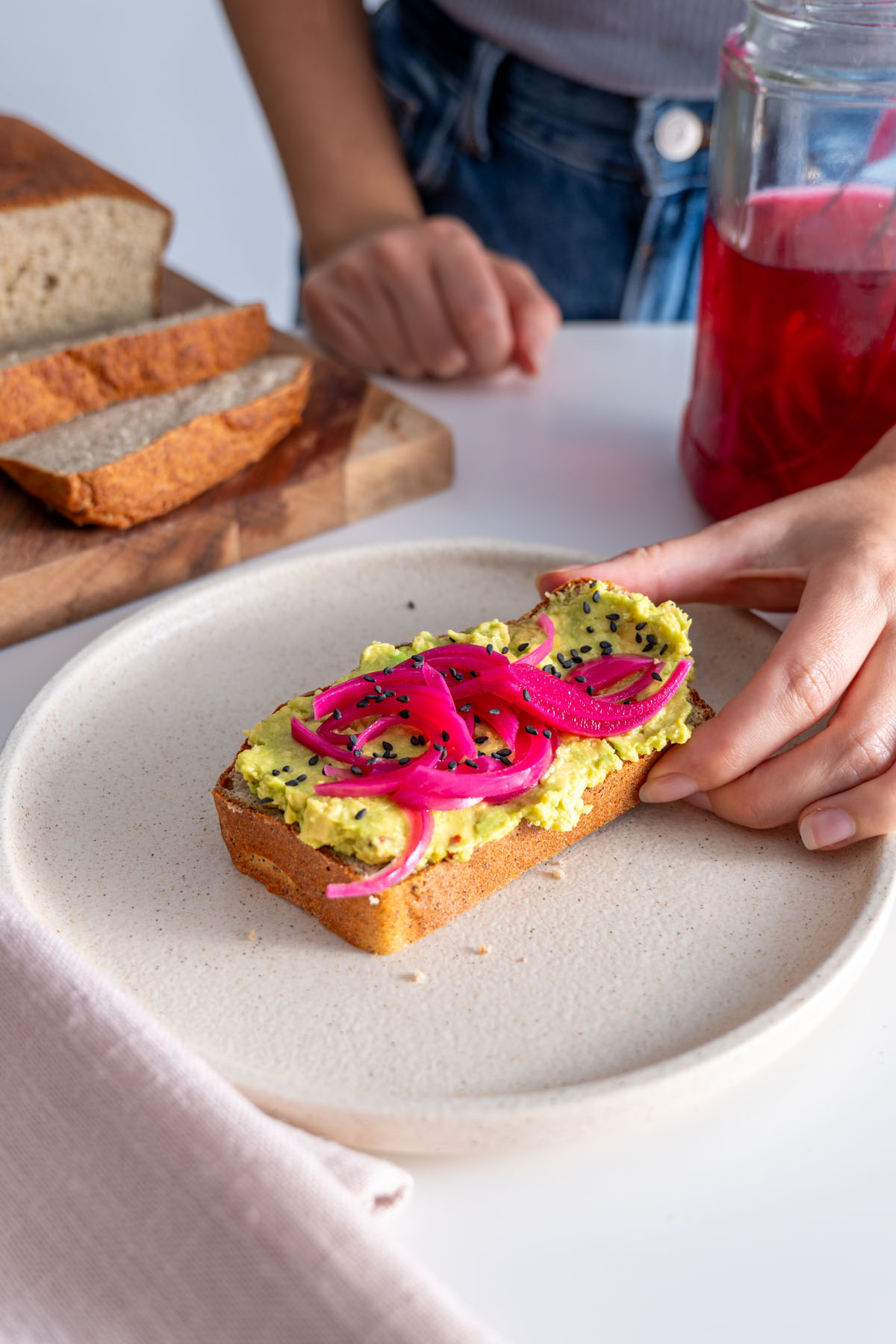 Sarah Cobacho reaching for a slice of red lentil quinoa bread with avocado spread and pickled onions on a beige plate.