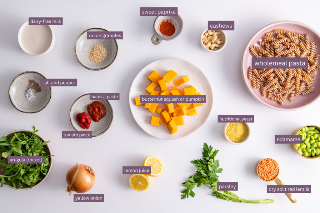 Vibrant ingredients for high protein pumpkin pasta on a white surface including pumpkin, pasta, and cashews