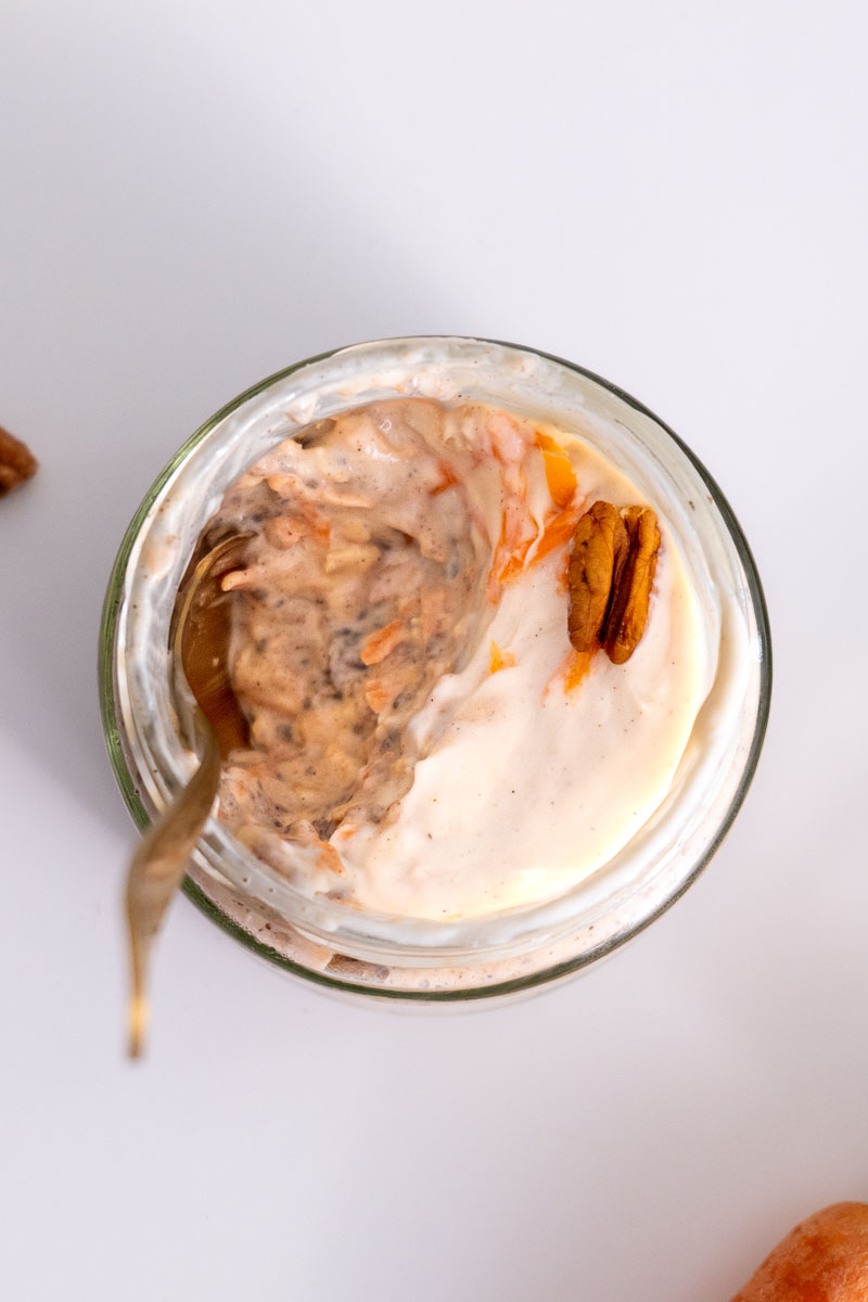 Top view of a jar of carrot cake overnight oats with a spoon inside, showing creamy layers and pecans on top.