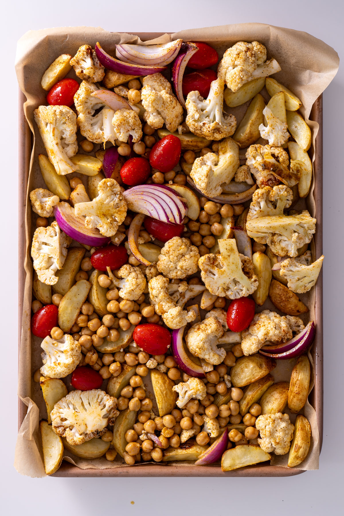 Chickpeas, cauliflower, potatoes, red onion, and spices arranged on a baking sheet ready to roast.