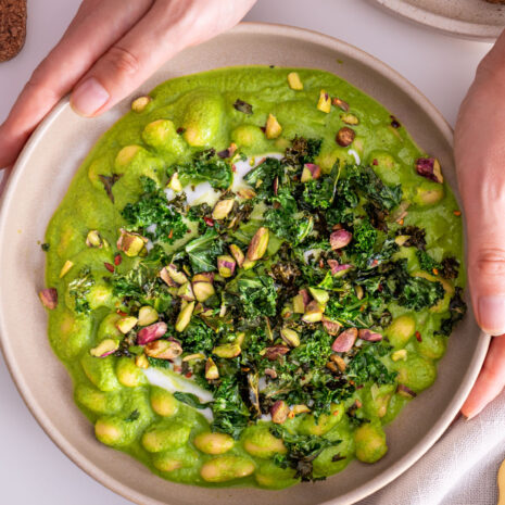 Super Green Butter Beans in a bowl, topped with crispy kale and pistachios, held by Sarah Cobacho