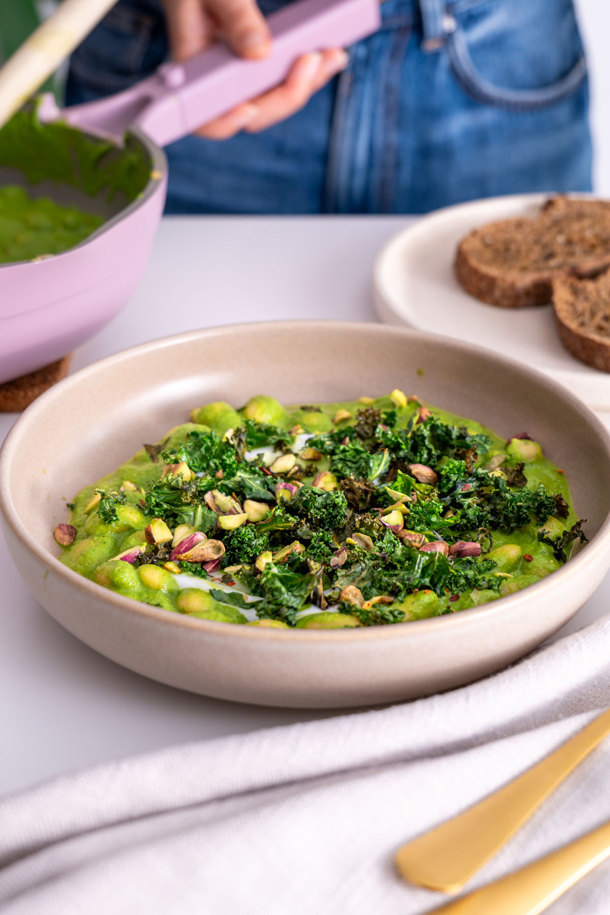 Super Green Butter Beans topped with crispy kale and pistachios on a white table with sourdough bread on the side