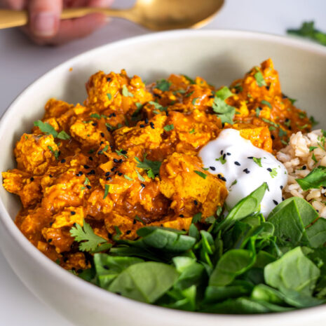 Sarah Cobacho holding a golden spoon while enjoying vegan butter chicken with tofu, served alongside fresh spinach and rice.
