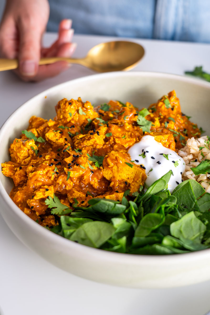 Sarah Cobacho holding a golden spoon while enjoying vegan butter chicken with tofu, served alongside fresh spinach and rice.