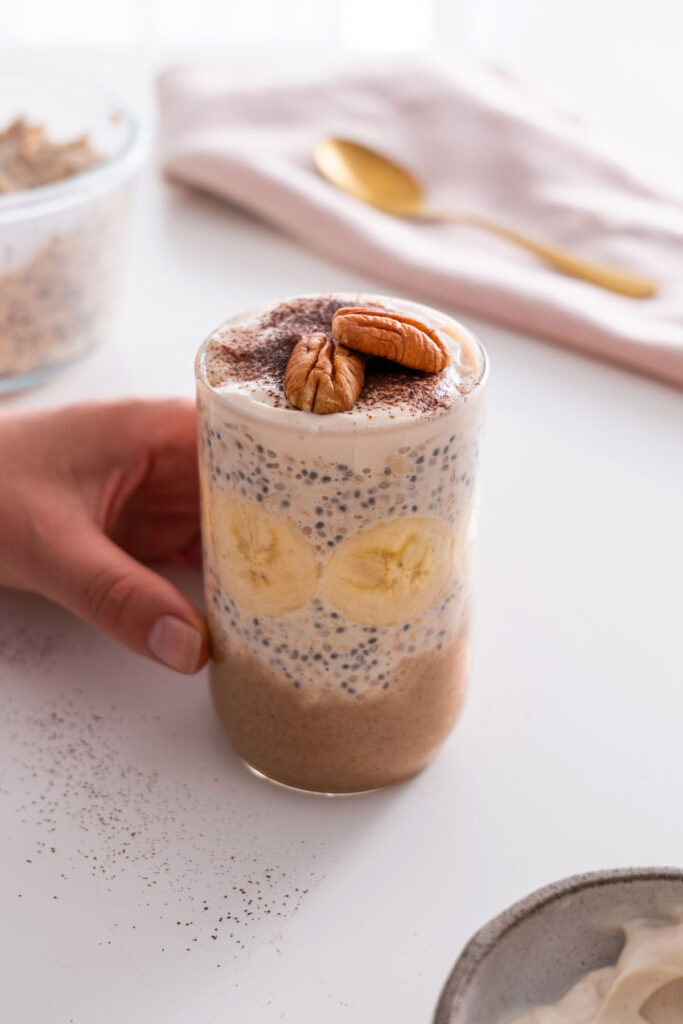 Banoffee Overnight Oats in a clear glass with banana slices, topped with cacao powder and pecans.