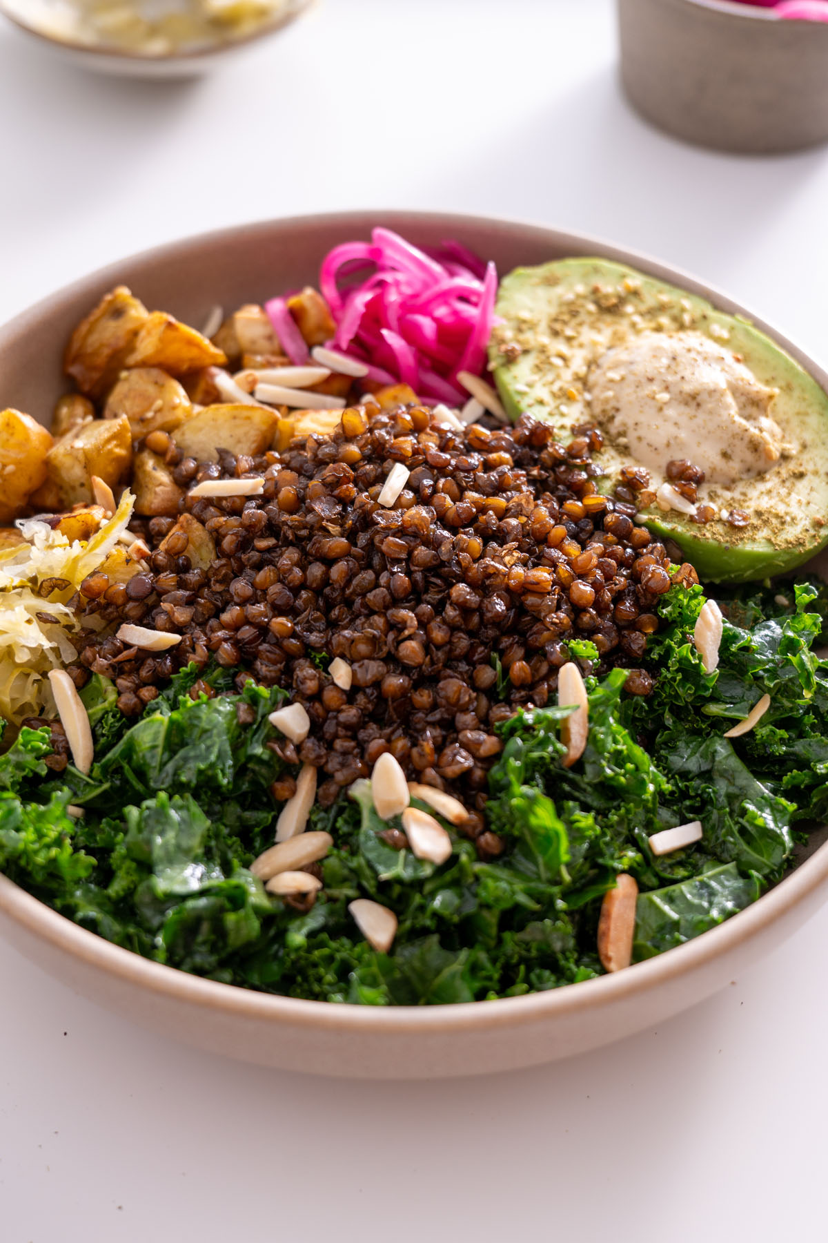 Crispy lentil and potato bowl assembled with kale, avocado, hummus, pickled onions, and almonds.