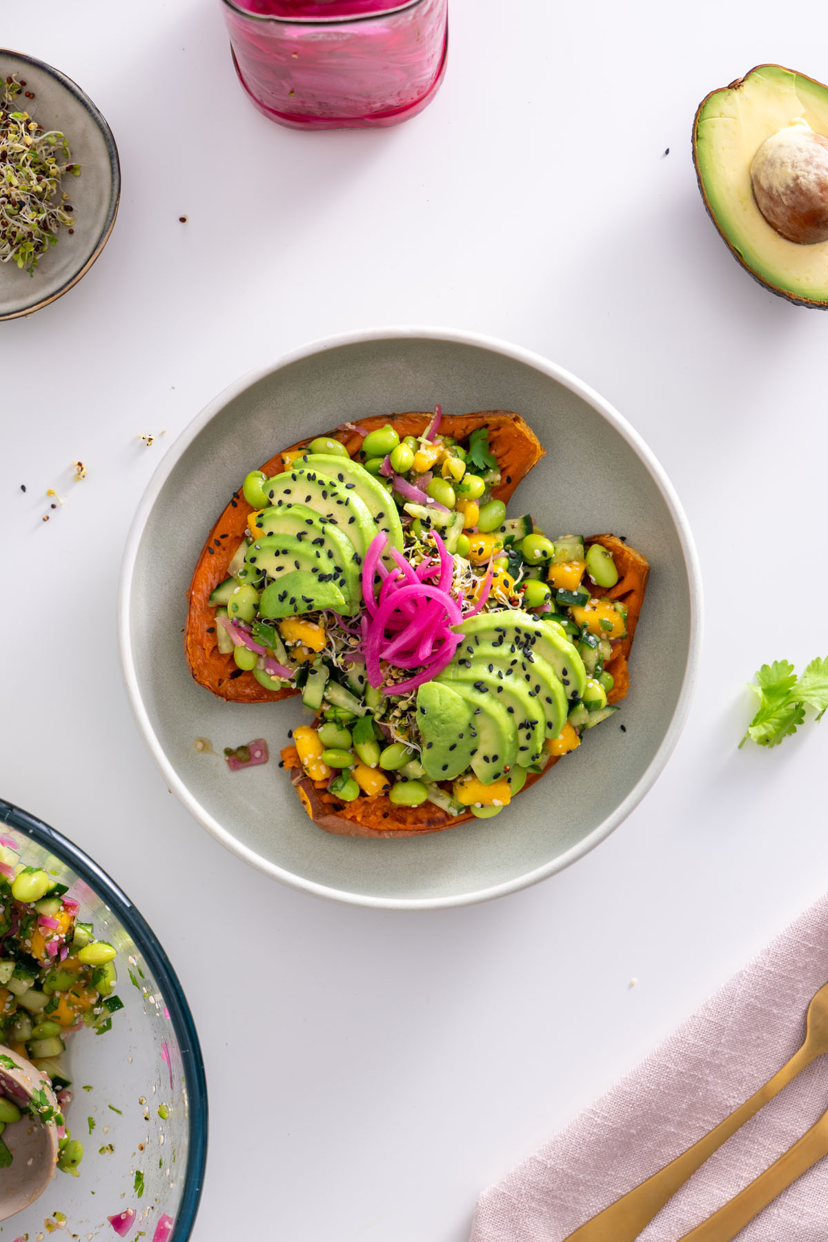Bowl with roasted sweet potato halves topped with edamame, mango, cucumber, avocado slices, and pickled onions.