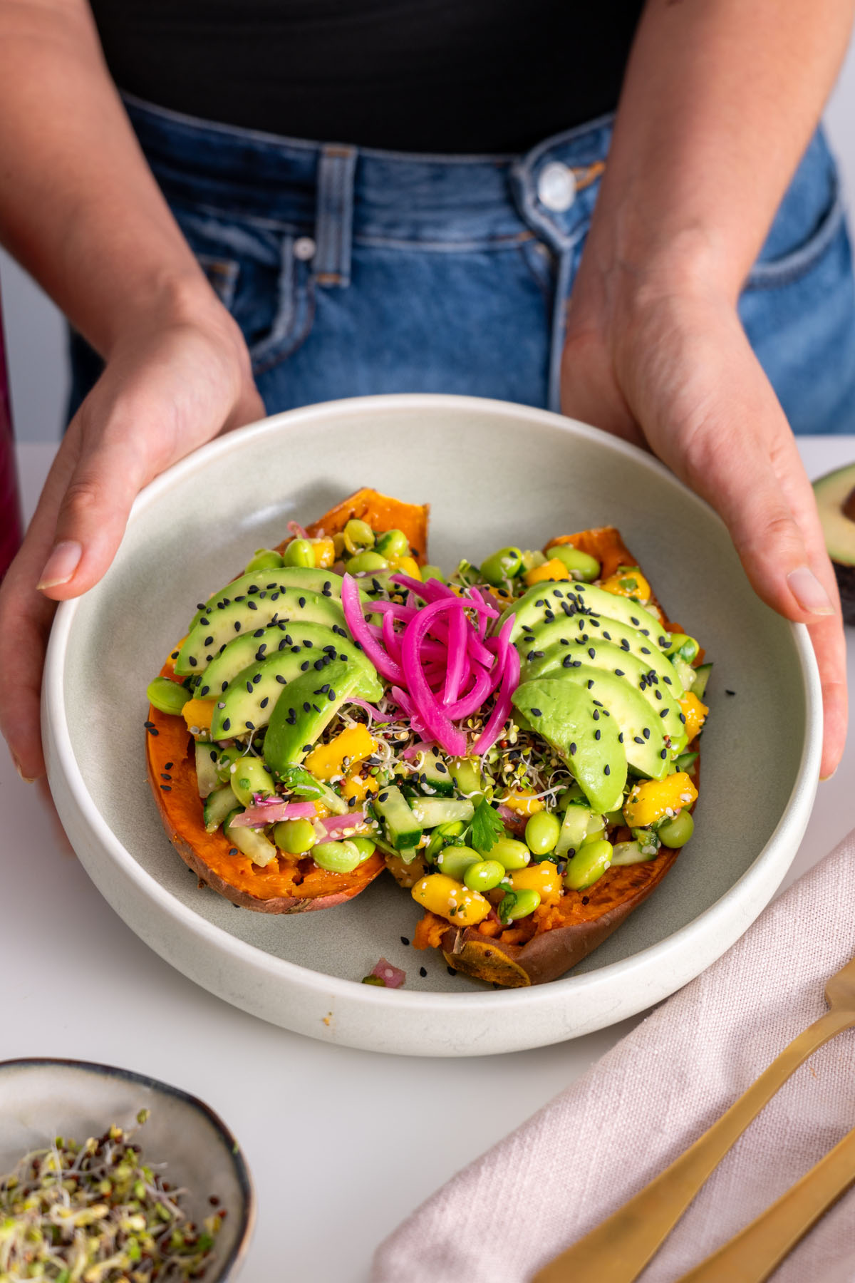 Sarah Cobacho holding a bowl of Edamame Sweet Potato Bowl with vibrant toppings and pickled onions in a light, fresh setting.