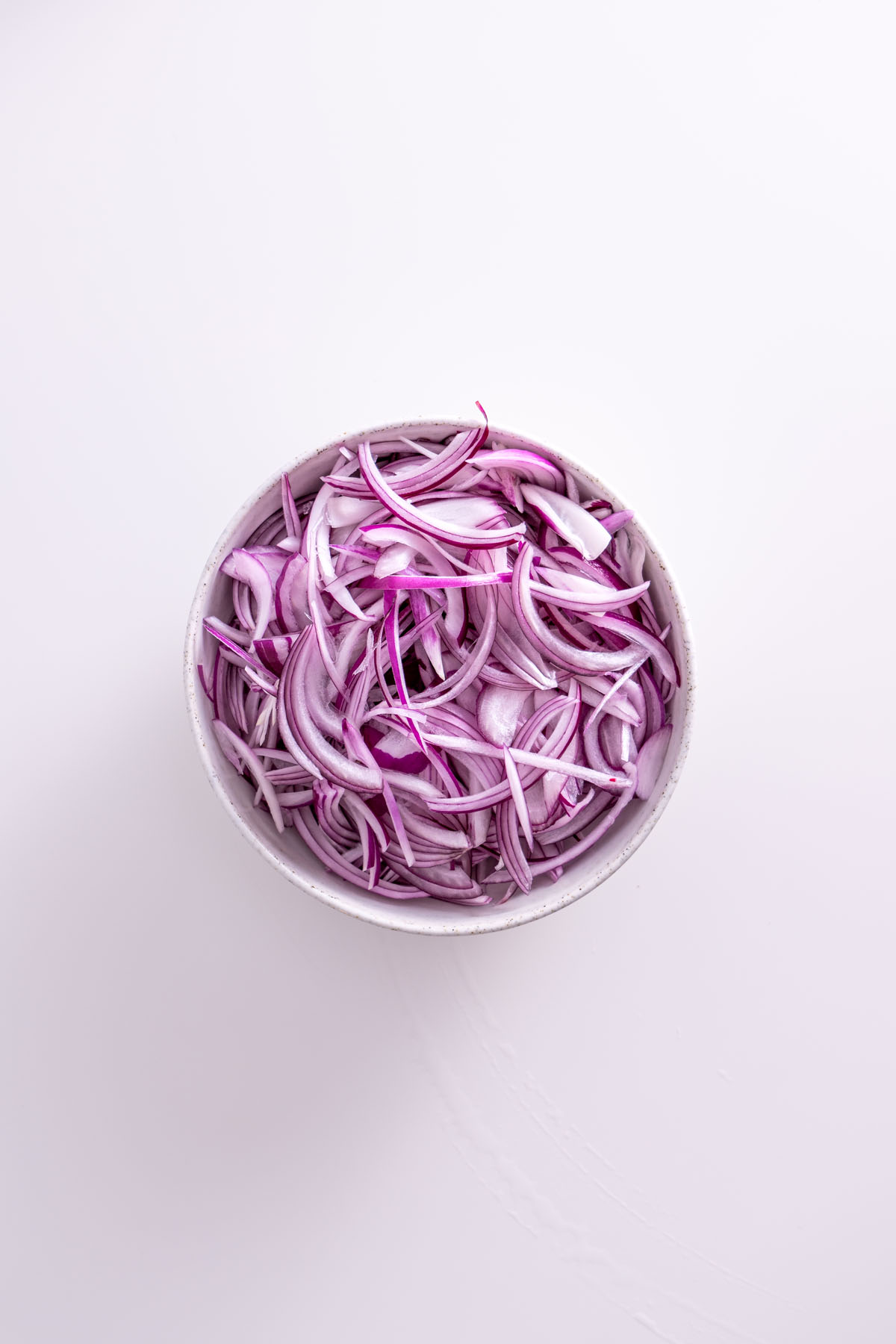 Thinly sliced red onions in a white bowl on a white surface.