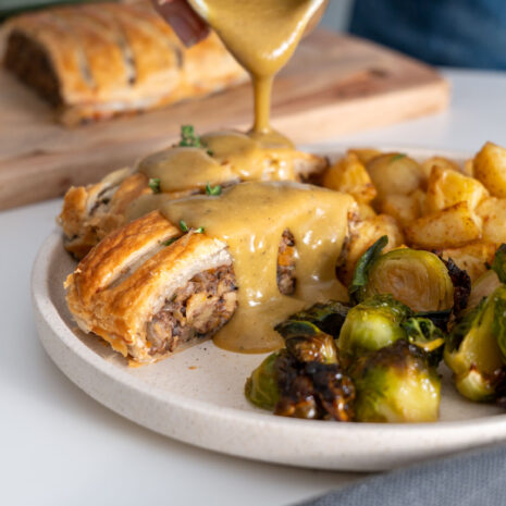 Sarah Cobacho pouring vegetable gravy over vegan mushroom Wellington slices on a plate of roasted vegetables.