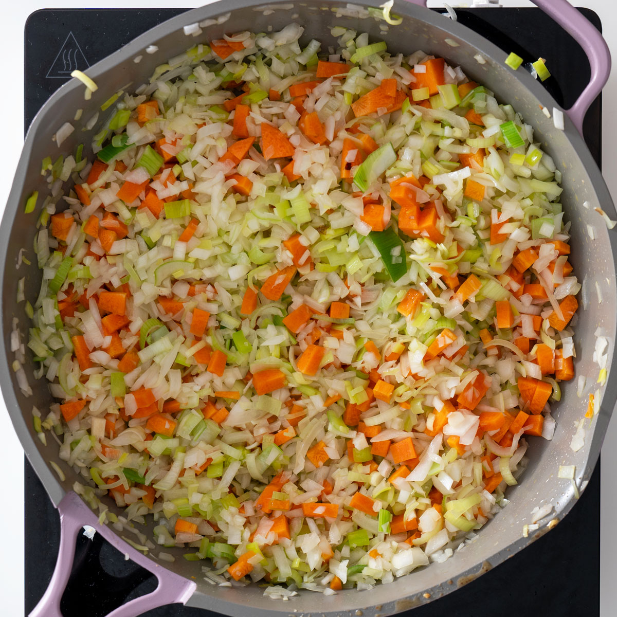 Process shot of chopped vegetables, including carrots, onions, and leeks, cooking in a large pan.