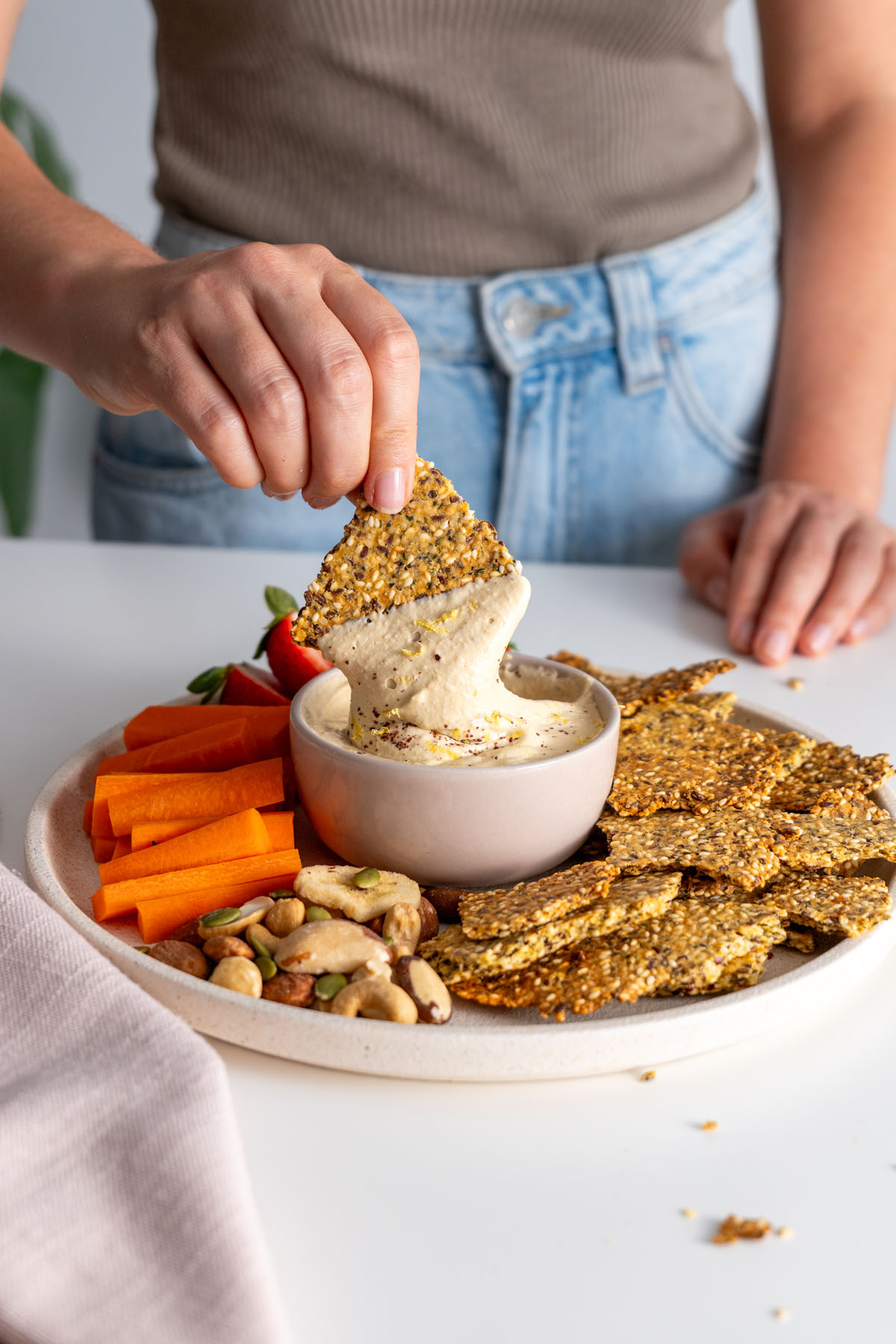 Sarah Cobacho holding a seeded cracker dipped in creamy dip with crackers and vegetable sticks on a serving plate.