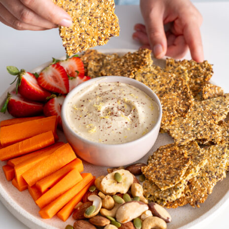 Sarah Cobacho holding a healthy seeded cracker above a platter with dip, strawberries, carrot sticks, and nuts.