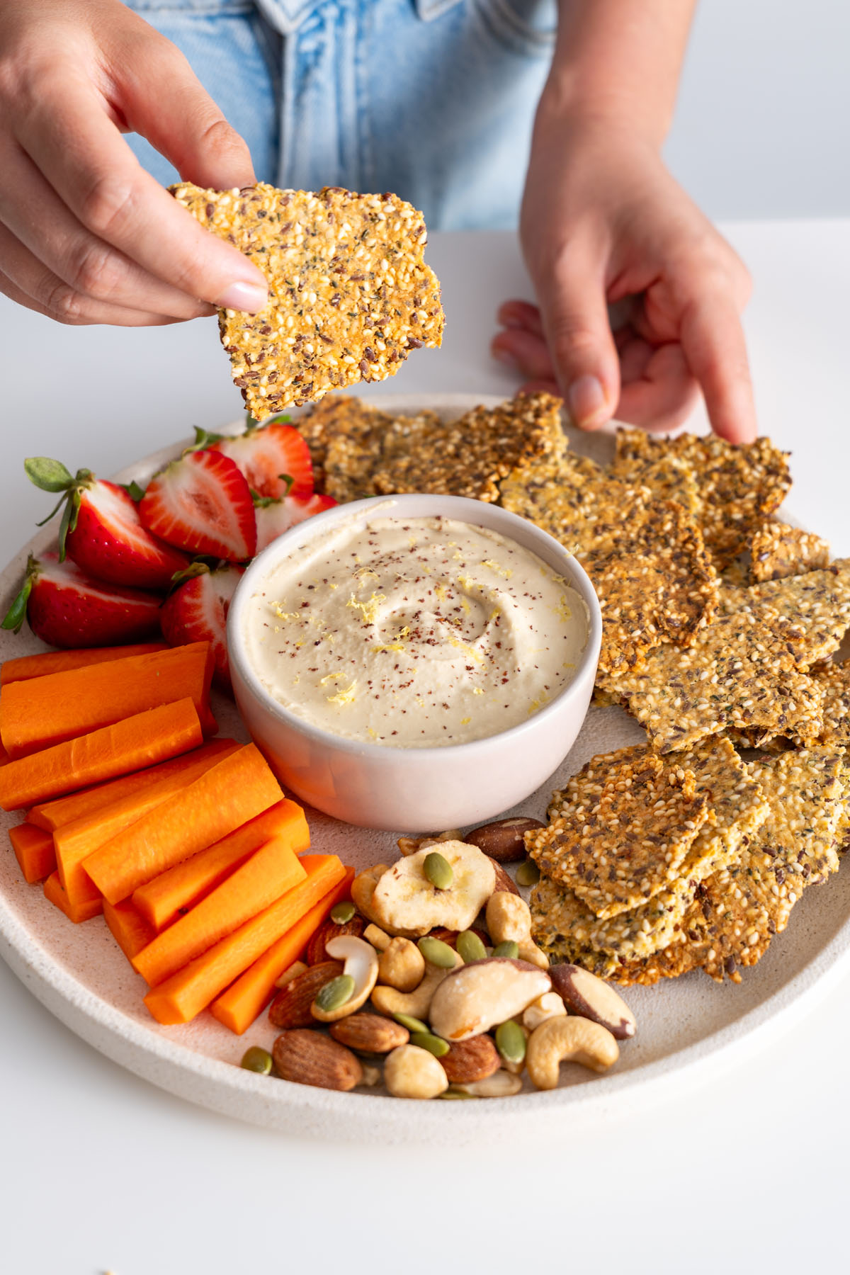Sarah Cobacho holding a healthy seeded cracker above a platter with dip, strawberries, carrot sticks, and nuts.