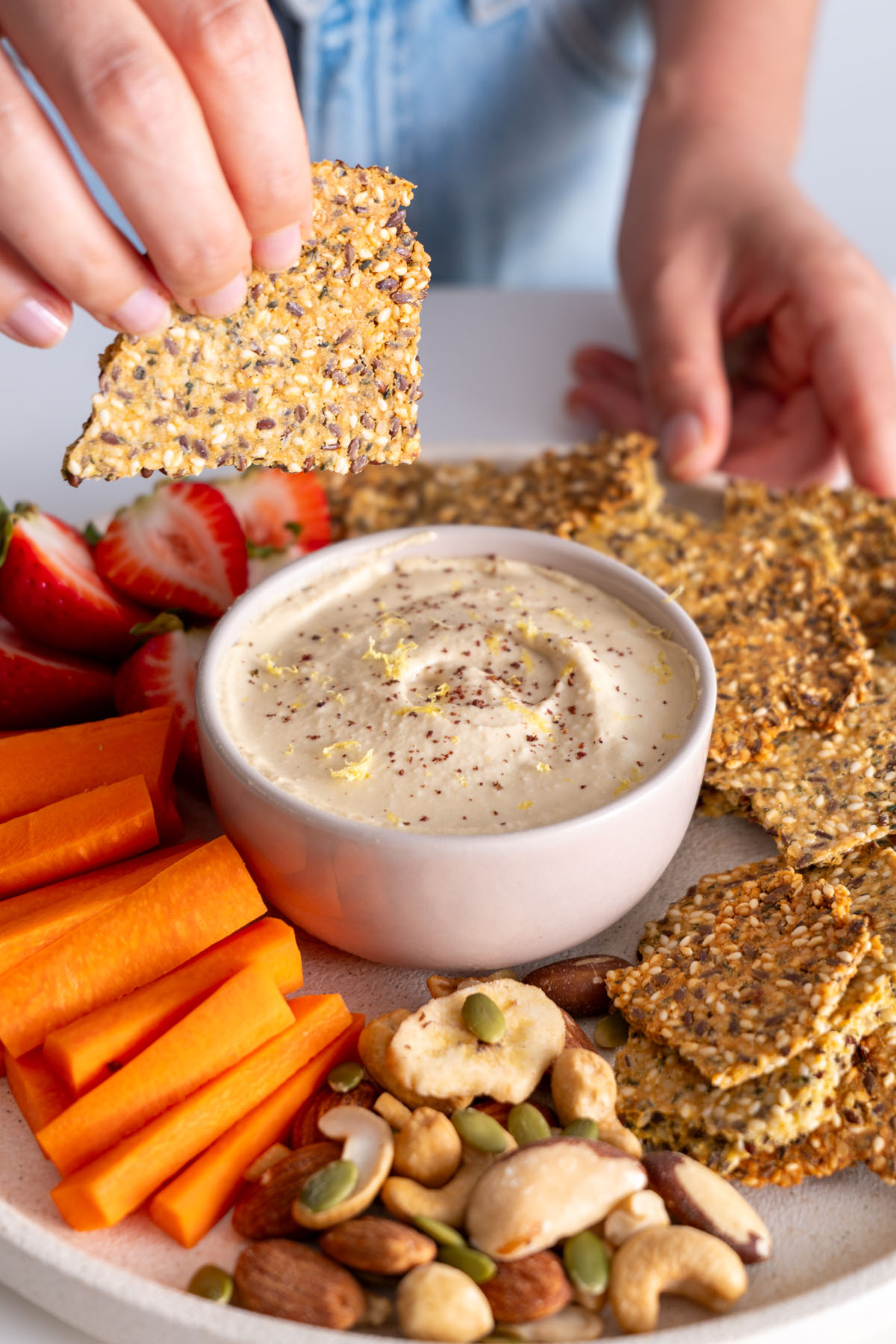 Sarah Cobacho holding a healthy seeded cracker close to a platter with dip, strawberries, carrot sticks, and nuts.
